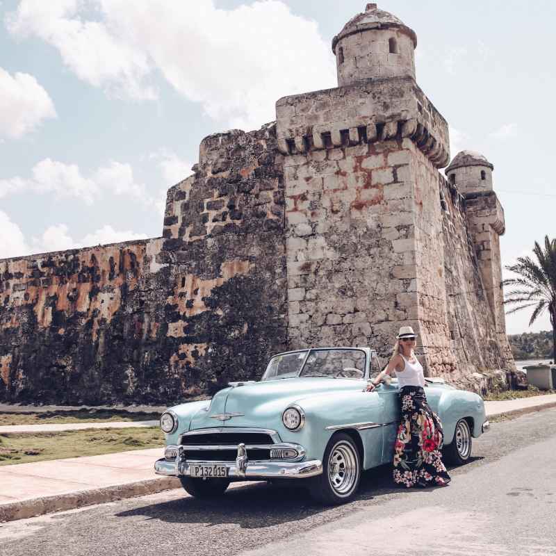 woman leaning against vintage old blue car with a stone tower in the background
